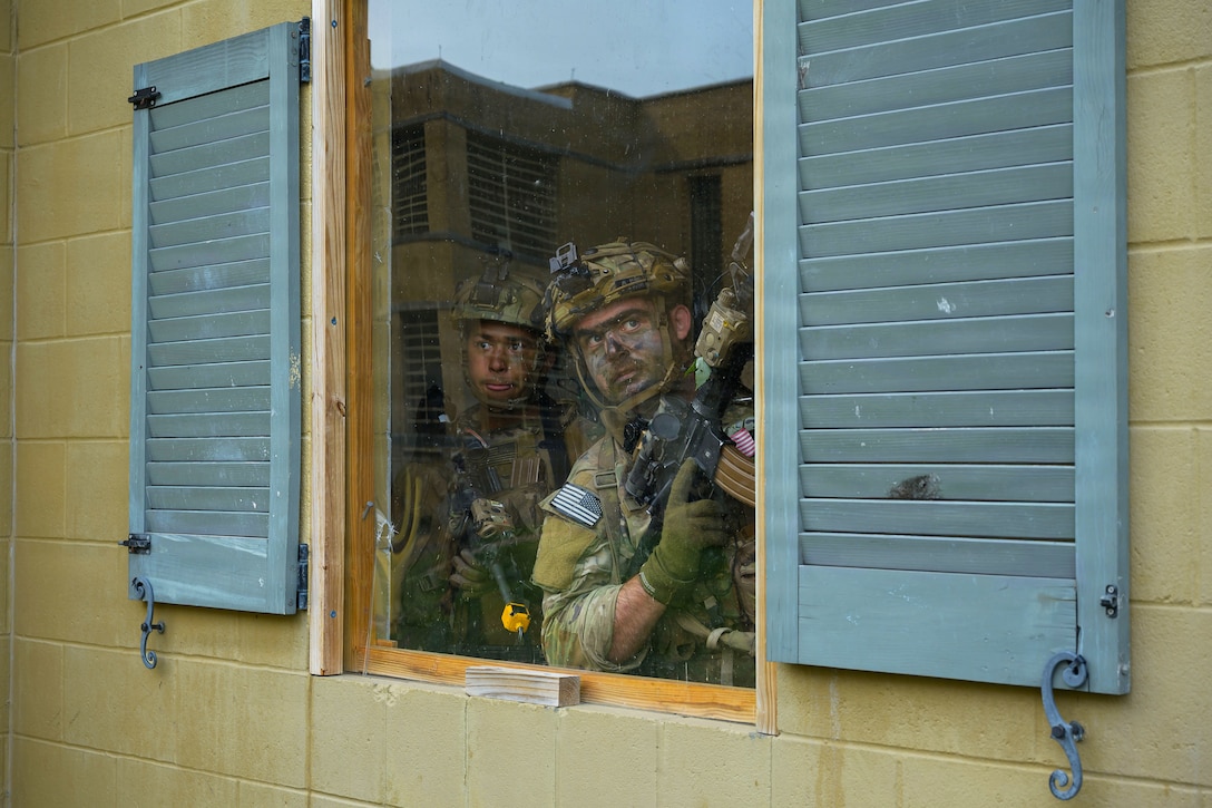 Two people in camouflage uniforms hold rifles and look out a window from a yellow house with green shutters. A building is reflected in the window's glass.