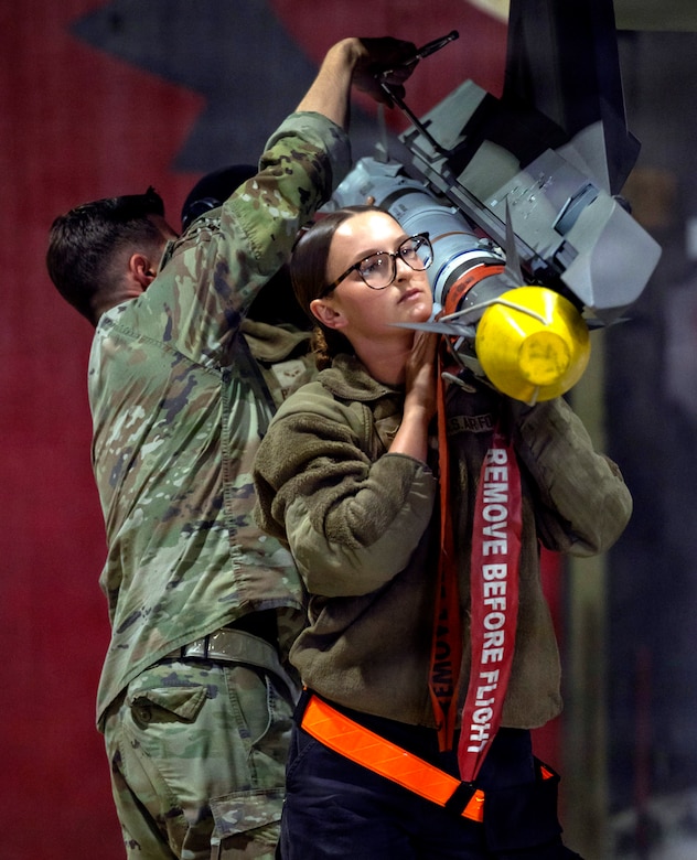 A woman wearing a military camouflage uniform lifts a missile on her shoulder as two men wearing similar attire connect it to a fighter jet.
