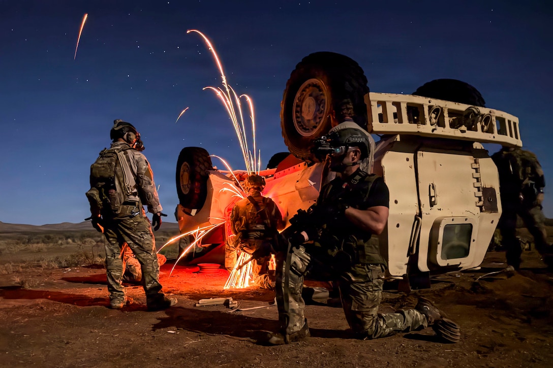 Four people in camouflage uniforms face an upside down military vehicle with flares shooting out of it while another person kneels on one knee on the ground facing the other direction to keep watch.