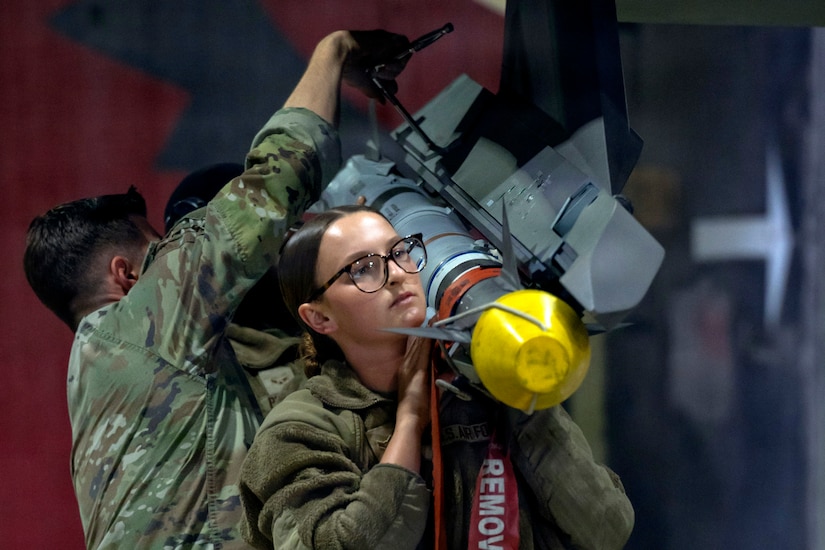 A woman wearing a military camouflage uniform lifts a missile on her shoulder as two men wearing similar attire connect it to a fighter jet.