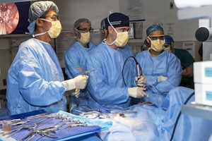 A man in medical scrubs performs surgery on a patient lying on a table as six other people in similar attire observe and assist.
