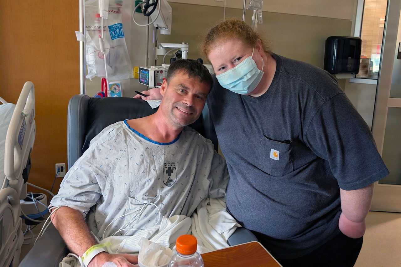 A man in a hospital gown poses for a photo with a woman in casual attire and a face mask while in a hospital room.