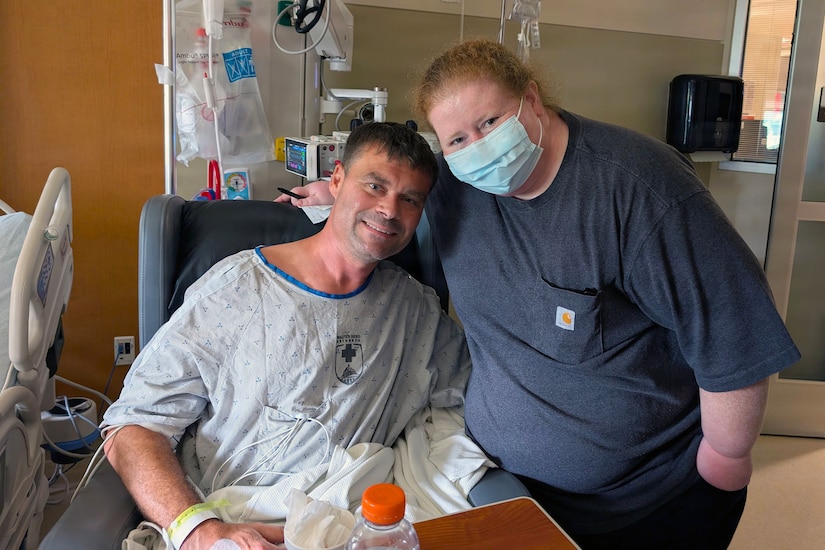 A man in a hospital gown poses for a photo with a woman in casual attire and a face mask while in a hospital room.