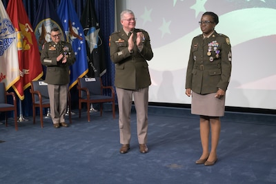 Three soldiers stand on a stage, with two of them applauding the third.