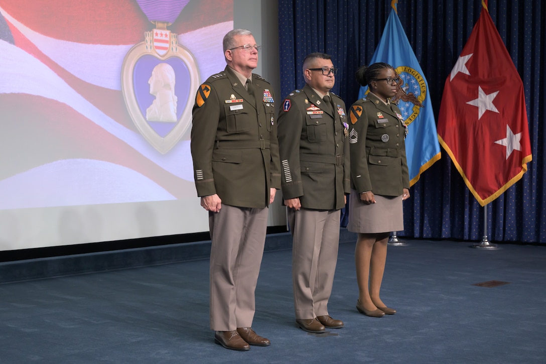 Three soldiers in Army Service Uniform stand at attention on a stage, with various flags behind them and the Purple Heart medal visible on the background screen.