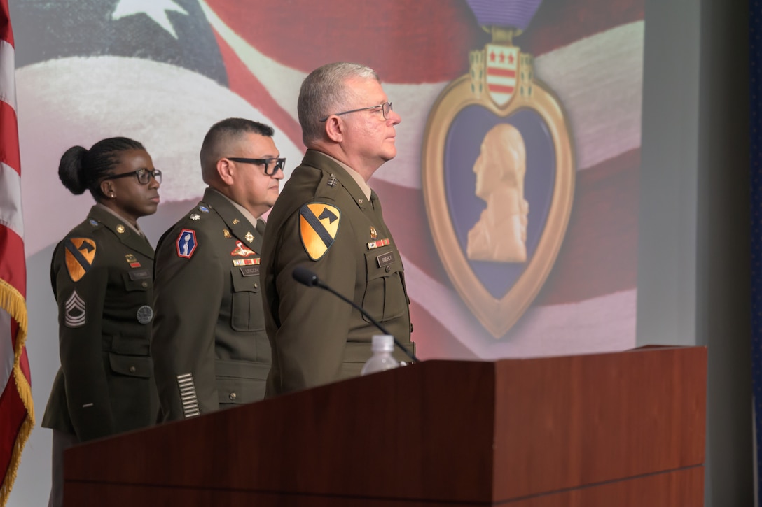 Three soldiers in Army Service Uniform stand at attention on a stage in front of a screen displaying a Purple Heart medal.