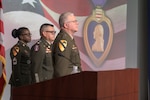 Three soldiers in Army Service Uniform stand at attention on a stage in front of a screen displaying a Purple Heart medal.