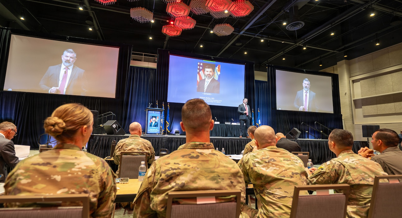 Man on stage in suit gives speech. Stands in front of his own photo.