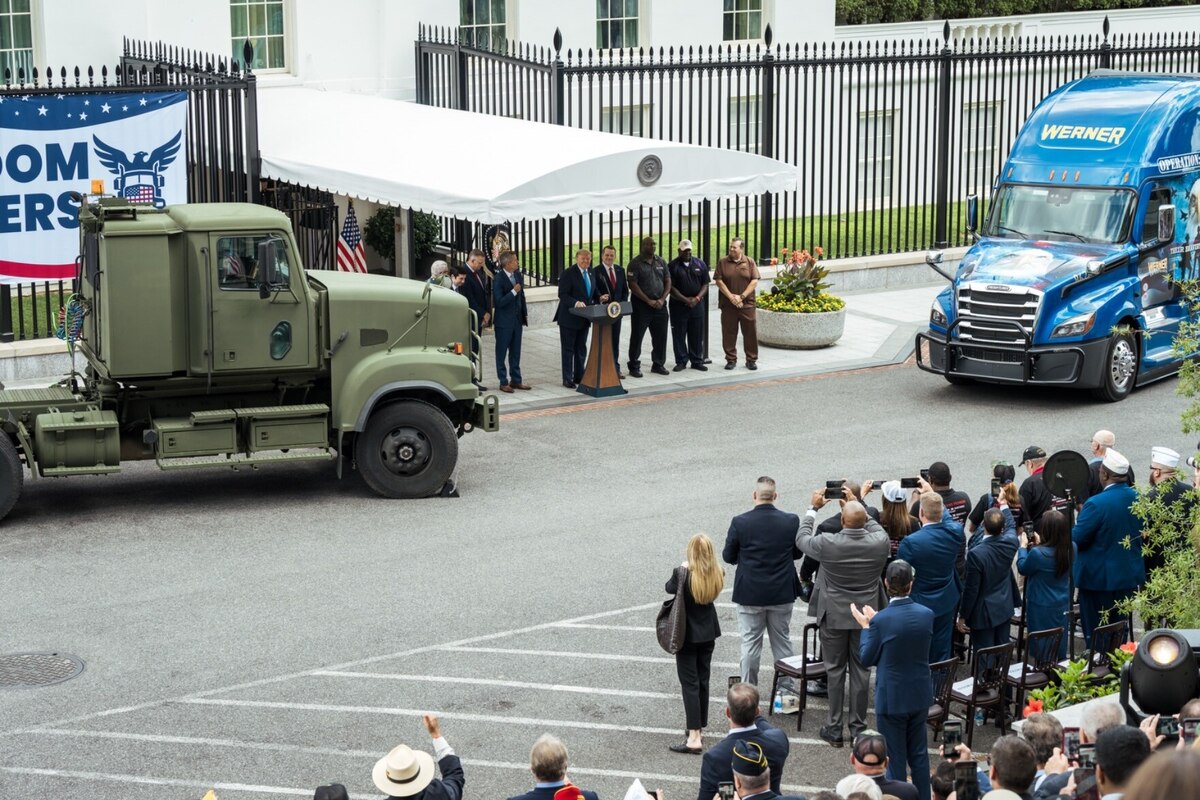 A man stands behind a lectern and is flanked on his left and right by several men and two large vehicles. In the rear is an entrance to the White House.