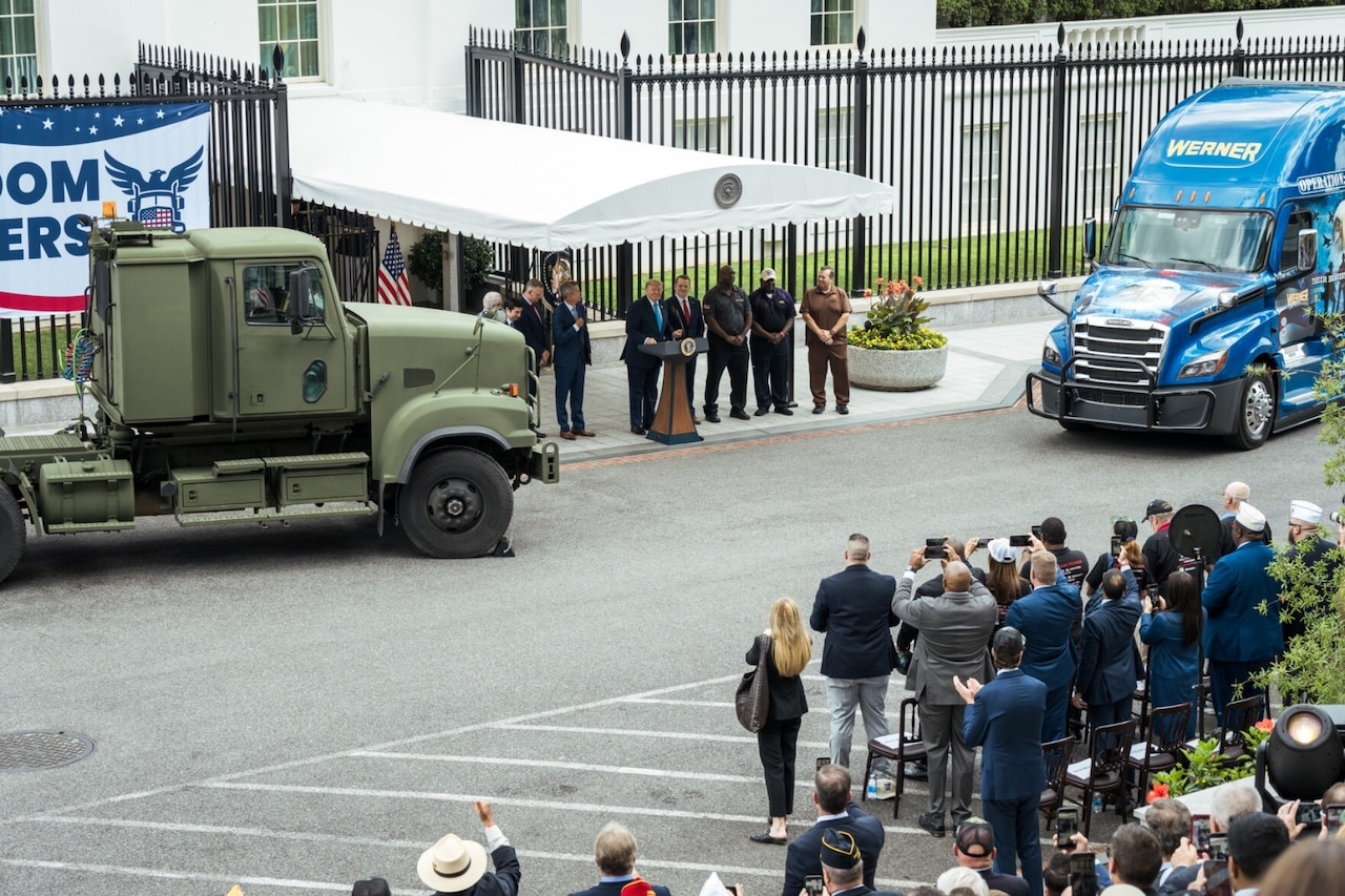 A man stands behind a lectern and is flanked on his left and right by several men and two large vehicles. In the rear is an entrance to the White House.