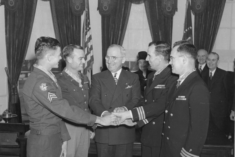 Four men wearing formal military uniforms and wearing medals around their necks, form a circle and join hands with a man in business attire.