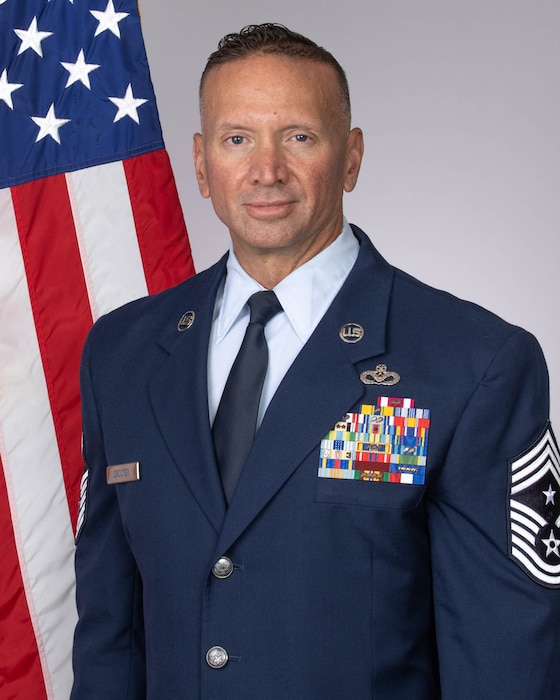 Male senior non-commissioned officer in dress blue Air Force uniform with American flag behind right shoulder