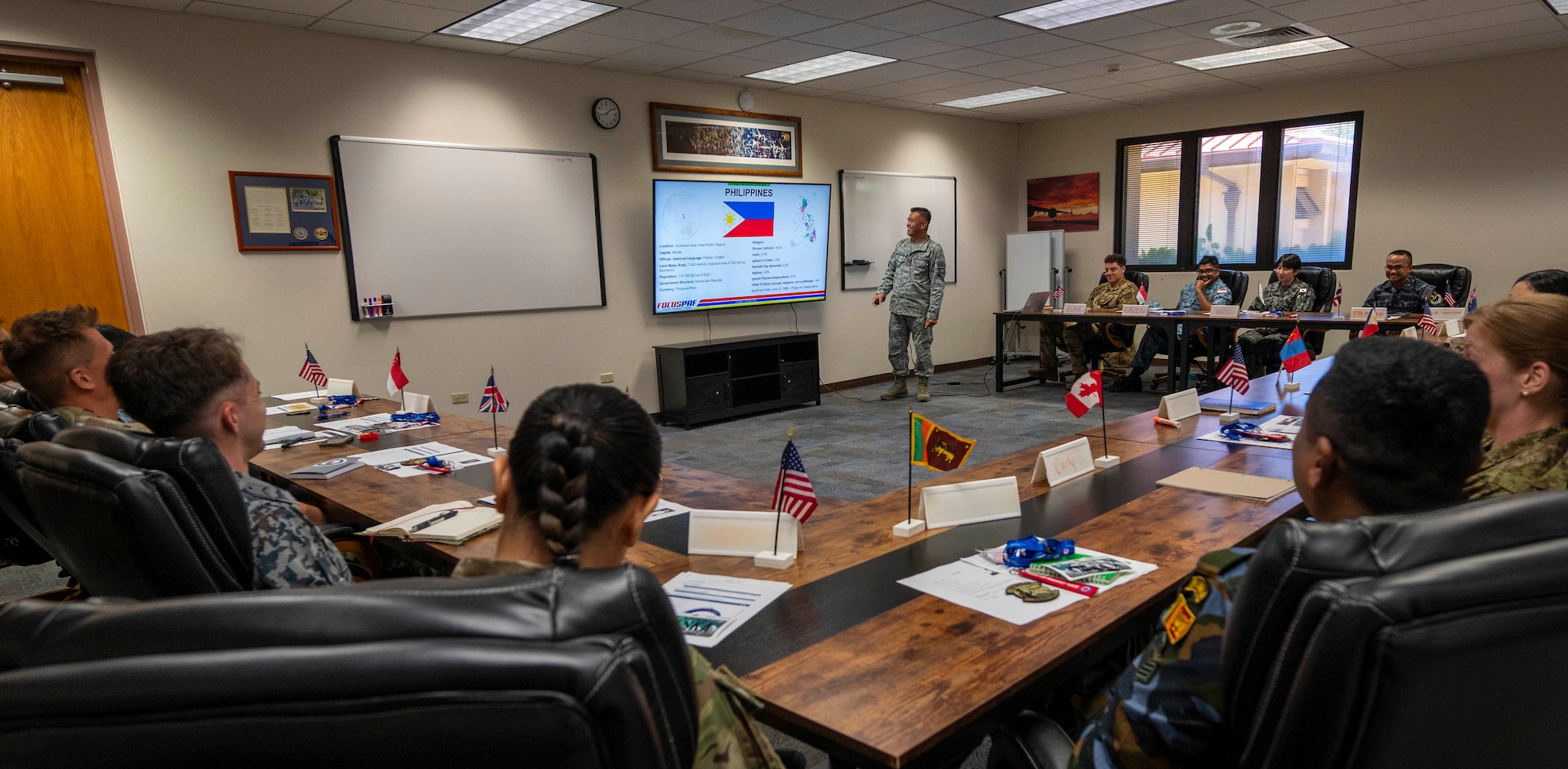 Classroom setting, a single military member is standing next to a TV giving a presentation to a room full of multinational military members.