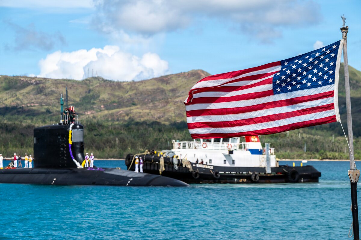 An American flag flies as a tugboat guides a submarine in the background.