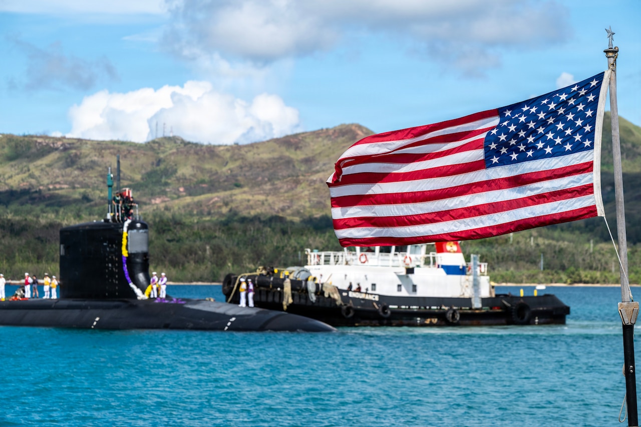 An American flag flies as a tugboat guides a submarine in the background.