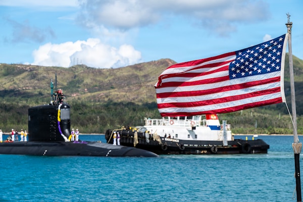 An American flag flies as a tugboat guides a submarine in the background.