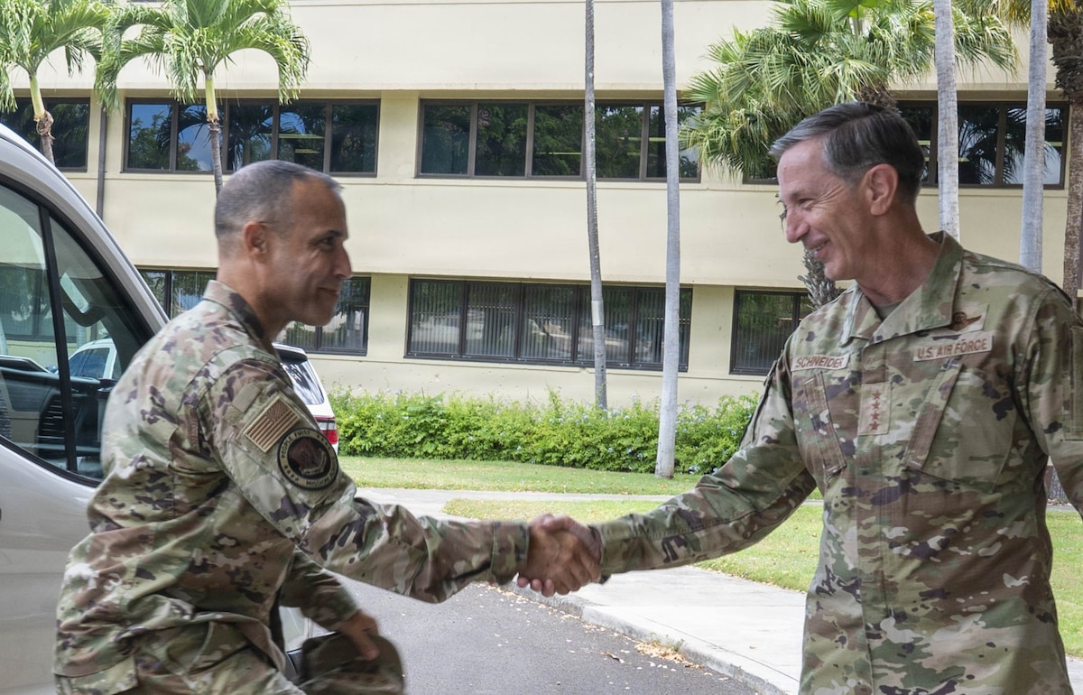 U.S. Air Force Gen. Kevin Schneider and Gen. Adrian Spain, both in camouflage uniforms, shake hands and smile at each other outdoors in front of a building with palm trees.