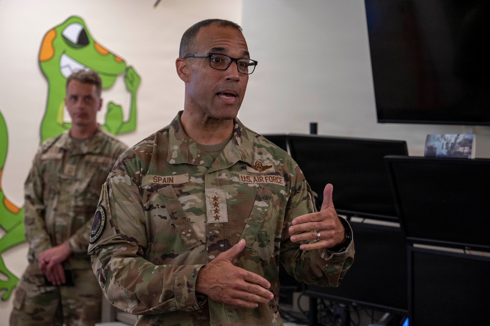 U.S. Air Force Gen. Adrian Spain, wearing glasses and a camouflage uniform, speaks and gestures with his hands while indoors. Another Airman stands in the background.