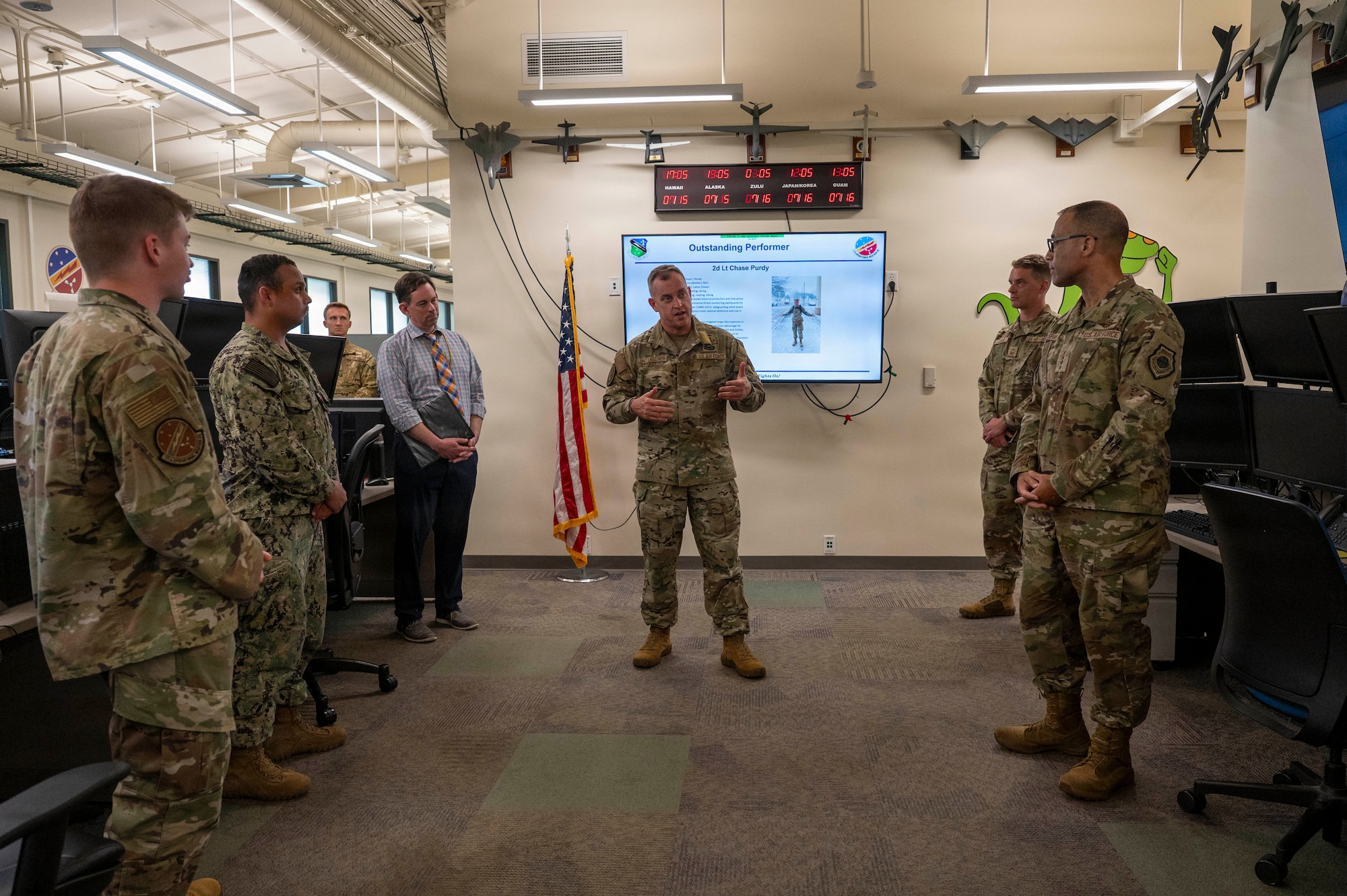 U.S. Air Force Chief Master Sgt. Jeremy Unterseher, in uniform, speaks to a group of service members gathered in an operations center. A presentation slide is visible on a screen behind him next to an American flag.