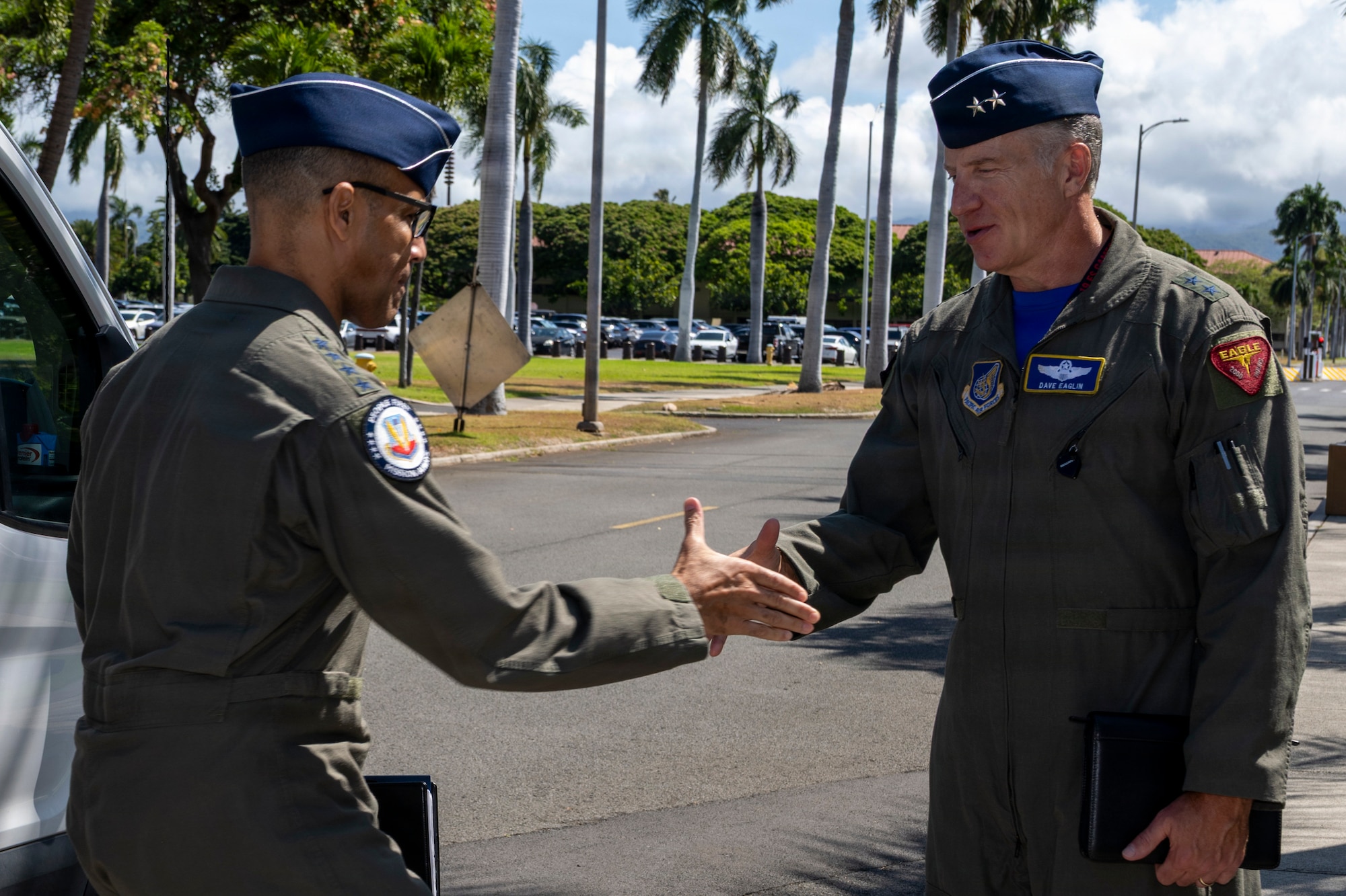 Two U.S. Air Force generals, Maj. Gen. Dave Eaglin and Gen. Adrian Spain, shake hands outdoors on a road lined with palm trees. Both are wearing green flight suits and blue garrison caps.