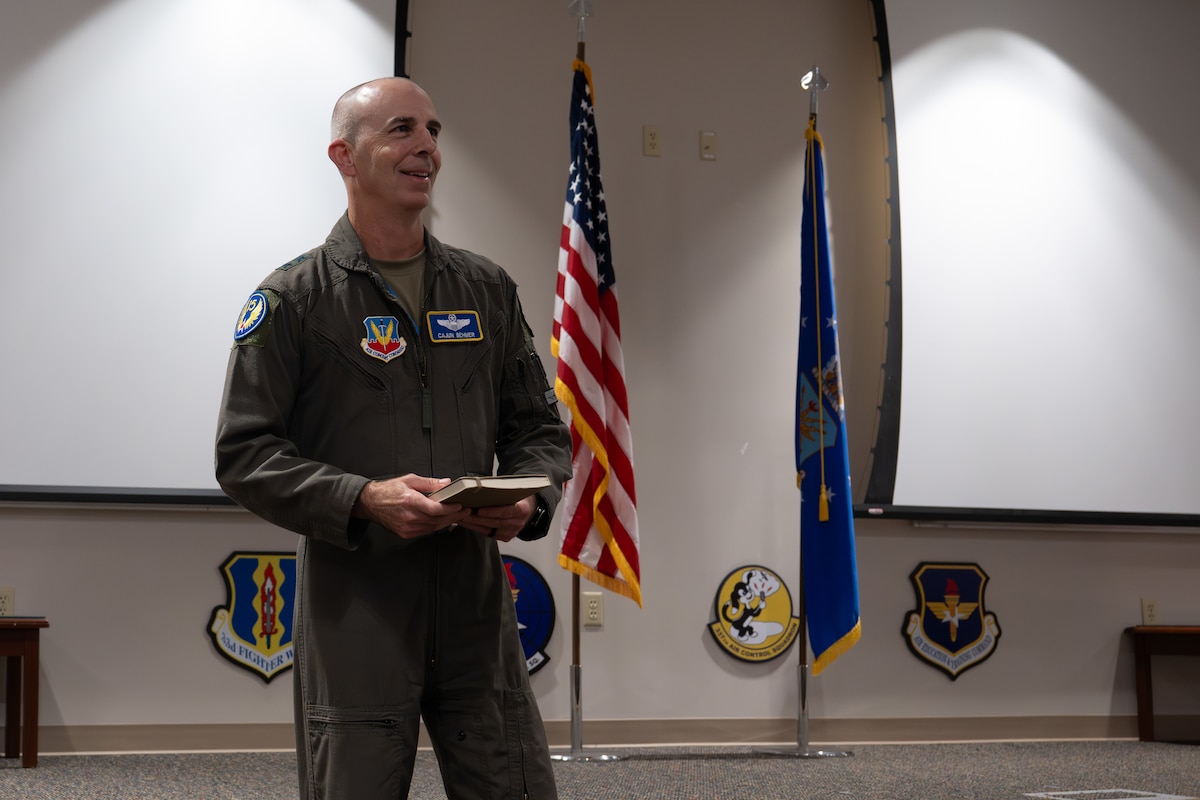 A U.S. Air Force pilot wearing a green flight suit stands on the left side of the frame, appearing to be in an auditorium. He is smiling and looking off frame