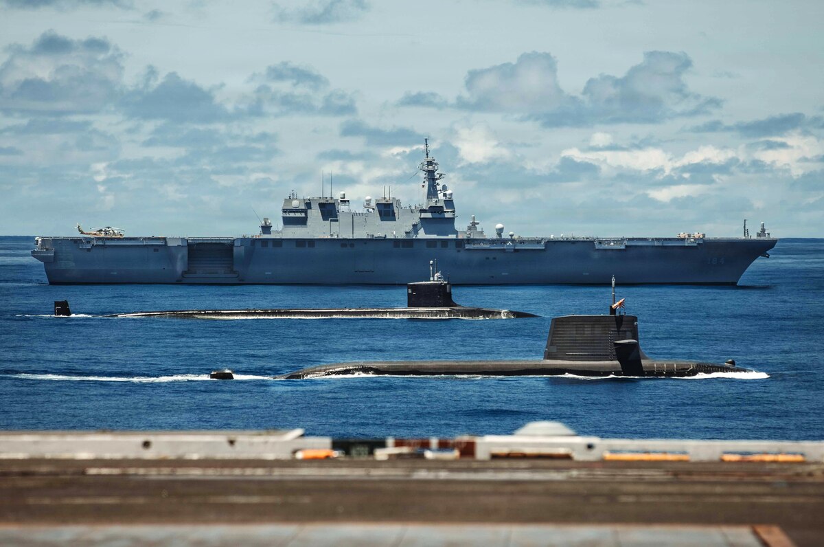 Two submarines and a large military ship sail in formation in the ocean.