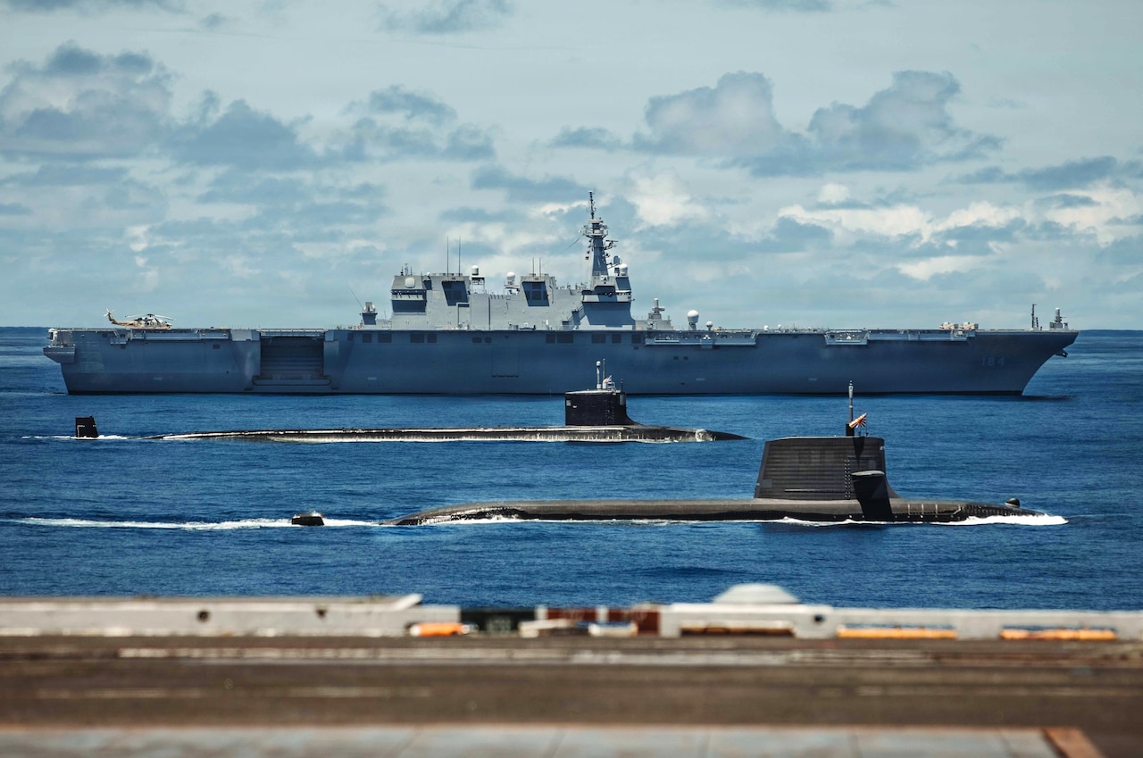Two submarines and a large military ship sail in formation in the ocean.
