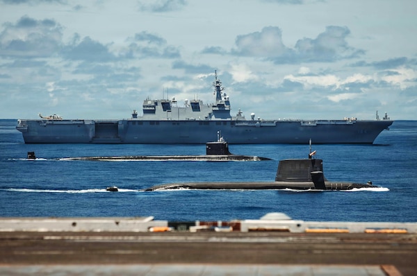 Two submarines and a large military ship sail in formation in the ocean.