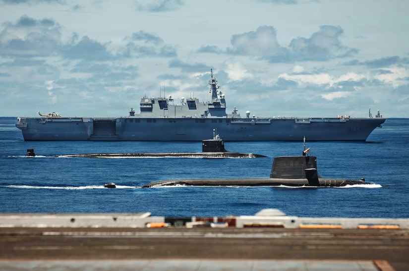Two submarines and a large military ship sail in formation in the ocean.