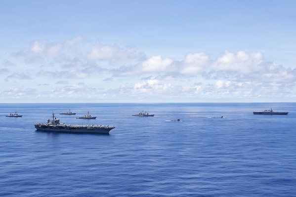 Six large military ships and two submarines sail in formation in the ocean.