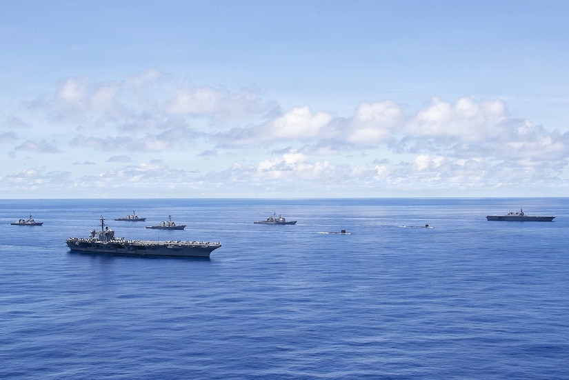 Six large military ships and two submarines sail in formation in the ocean.