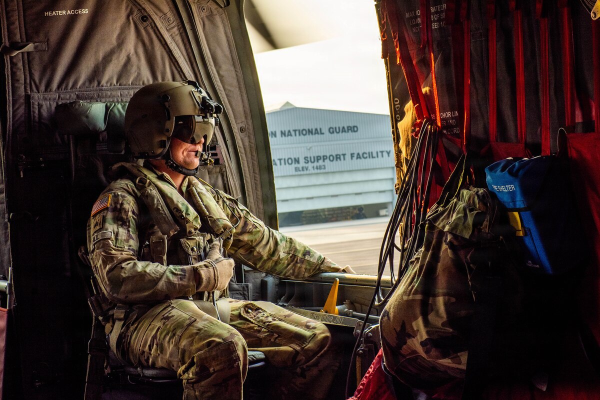 A man wearing a military camouflage uniform and flight helmet sits inside of a helicopter as it prepares to lift off.