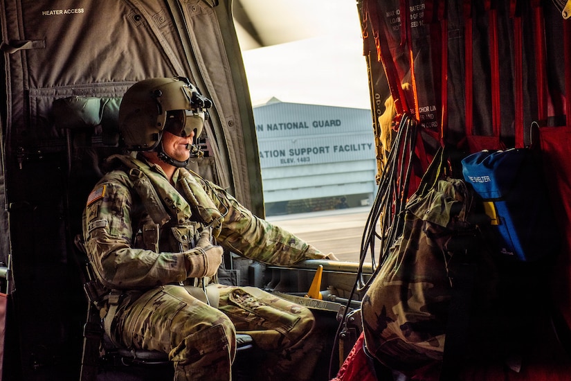 A man wearing a military camouflage uniform and flight helmet sits inside of a helicopter as it prepares to lift off.