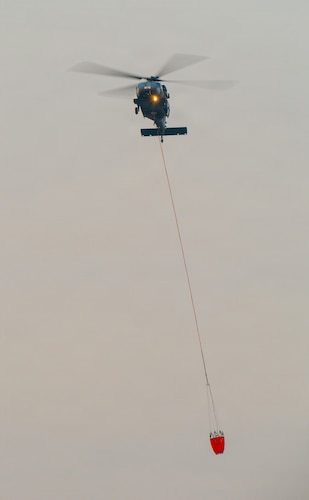 A helicopter flies through wildfire smoke carrying a water bucket connected to a cable.