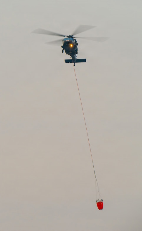 A helicopter flies through wildfire smoke carrying a water bucket connected to a cable.