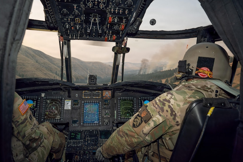 Two people wearing military camouflage uniforms and flight helmets sit in the cockpit of a helicopter while flying toward a wildfire.