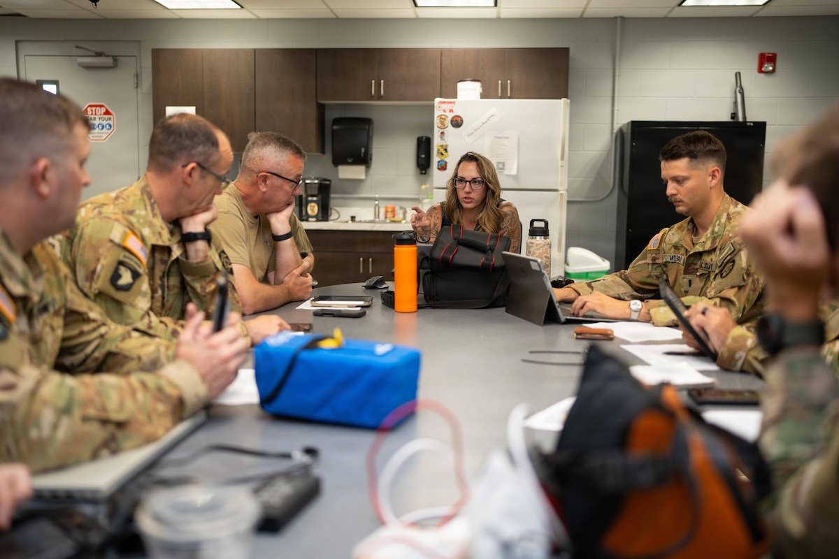 A woman sits at the head of a table in a meeting room and briefs people dressed in military camouflage uniforms sitting to the left and right of her.