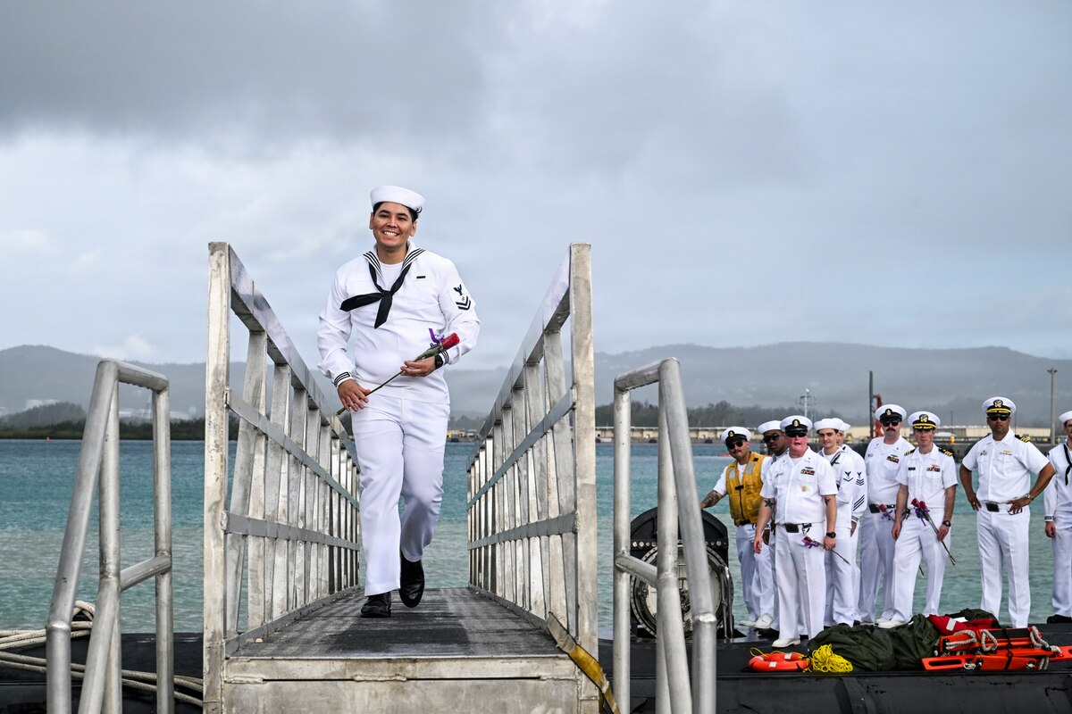 A sailor in a military dress uniform walks across a bridge from a submarine as several people in similar attire stand on top of the vessel in the background.