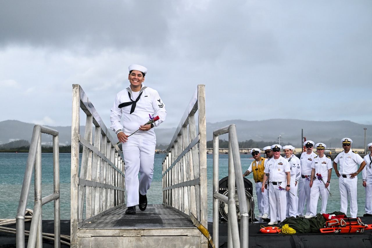 A sailor in a military dress uniform walks across a bridge from a submarine as several people in similar attire stand on top of the vessel in the background.
