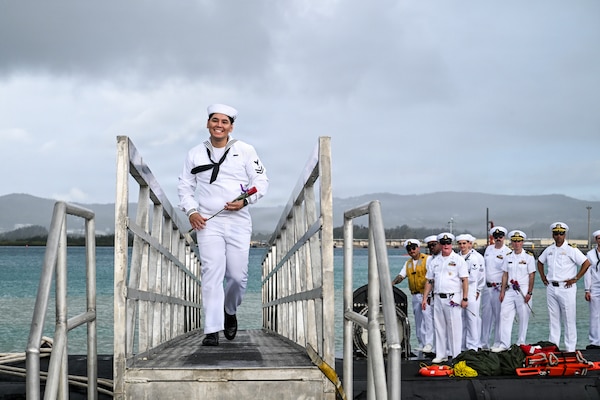 A sailor in a military dress uniform walks across a bridge from a submarine as several people in similar attire stand on top of the vessel in the background.