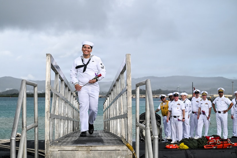 A sailor in a military dress uniform walks across a bridge from a submarine as several people in similar attire stand on top of the vessel in the background.