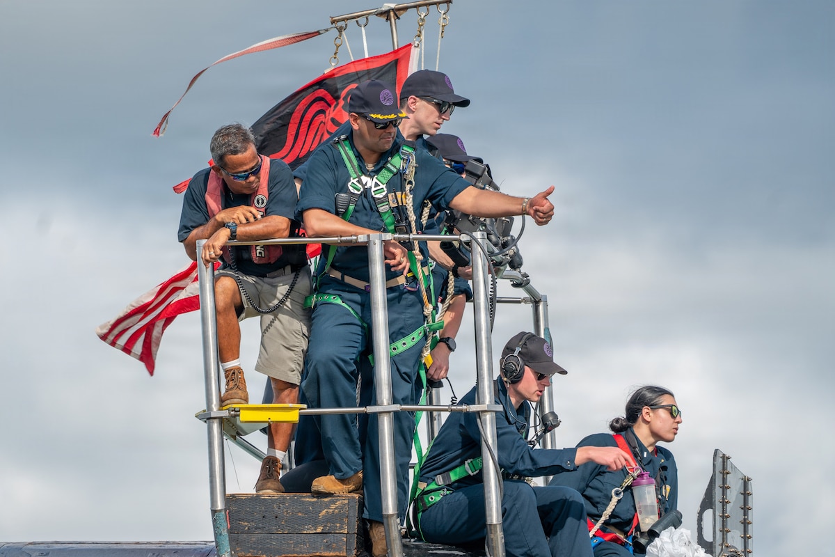 Six people in military uniforms and casual attire stand on top of a submarine as they signal to other people.