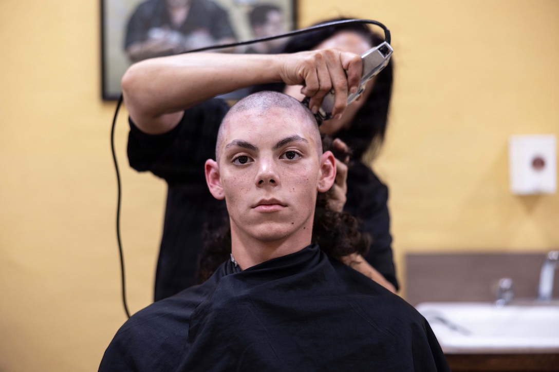 A seated Marine Corps recruit covered with a black cape gets a buzz cut from a barber standing against a blurred yellow wall.