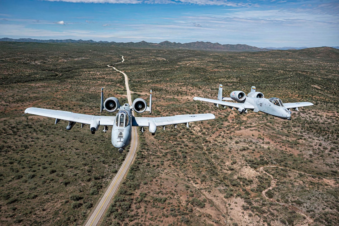 Two aircraft fly next to each other in a blue sky over a desert with mountains in the distance.