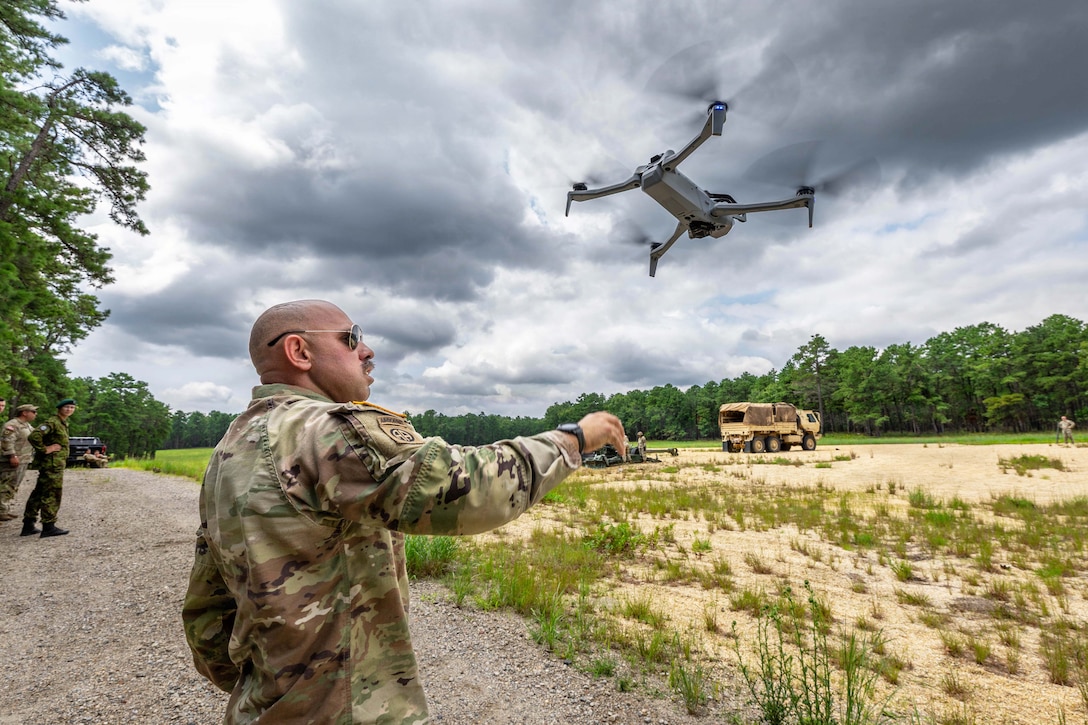 A soldier standing to the left launches a drone over a field toward a gray sky as other soldiers watch in the distance.
