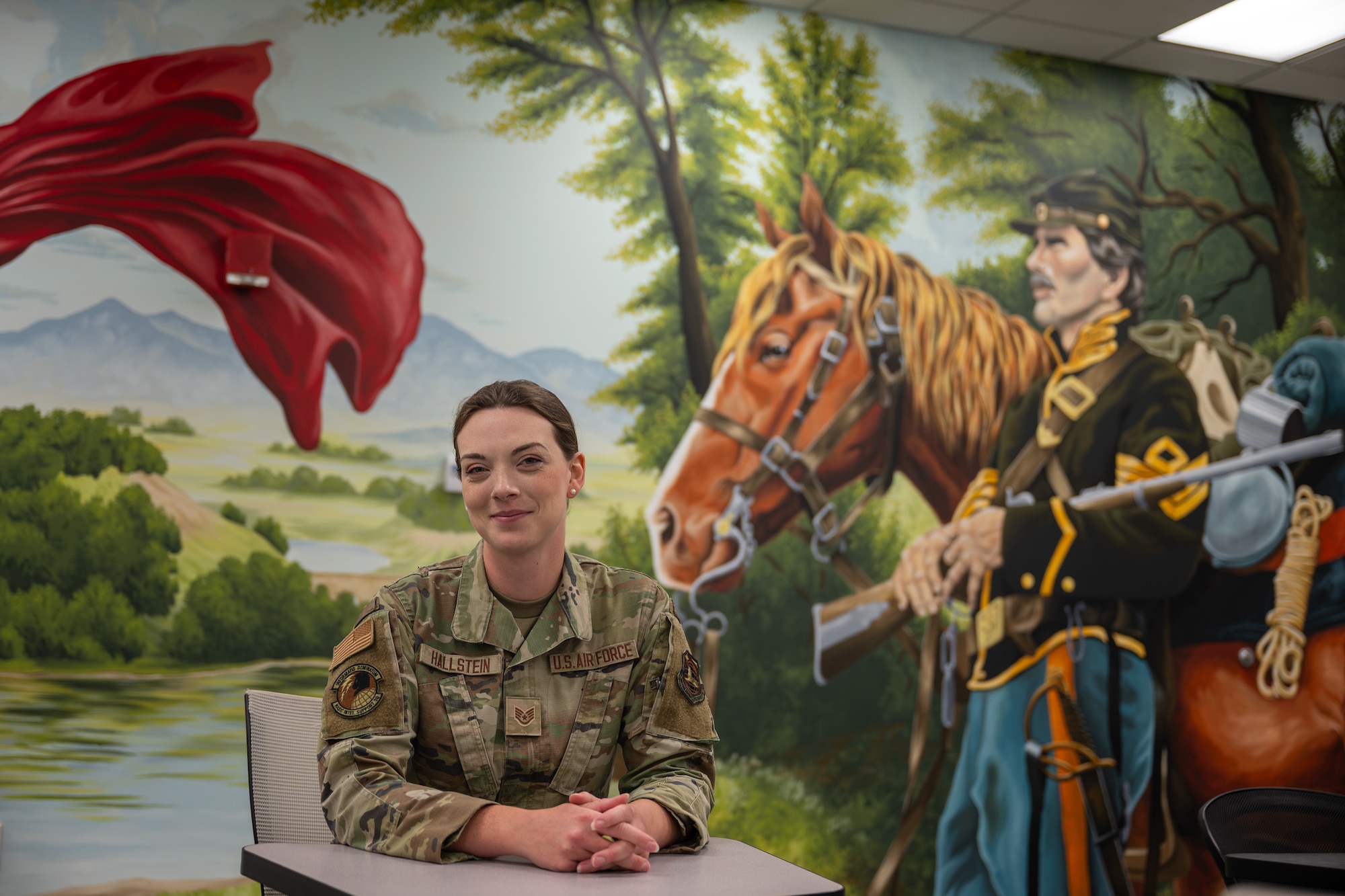 Staff Sgt. Sara Hallstein, Intelligence Support Squadron noncommissioned officer in charge, poses for a photo next to a mural she painted at Wright-Patterson Air Force Base, Ohio, June 25, 2026. Through her art, Hallstein hopes to encourage others to uncover their own "hidden gifts" and redefine how creativity fits into the broader National Air and Space Intelligence Center mission. (U.S. Air Force photo by Staff Sgt. Kristof J. Rixmann)