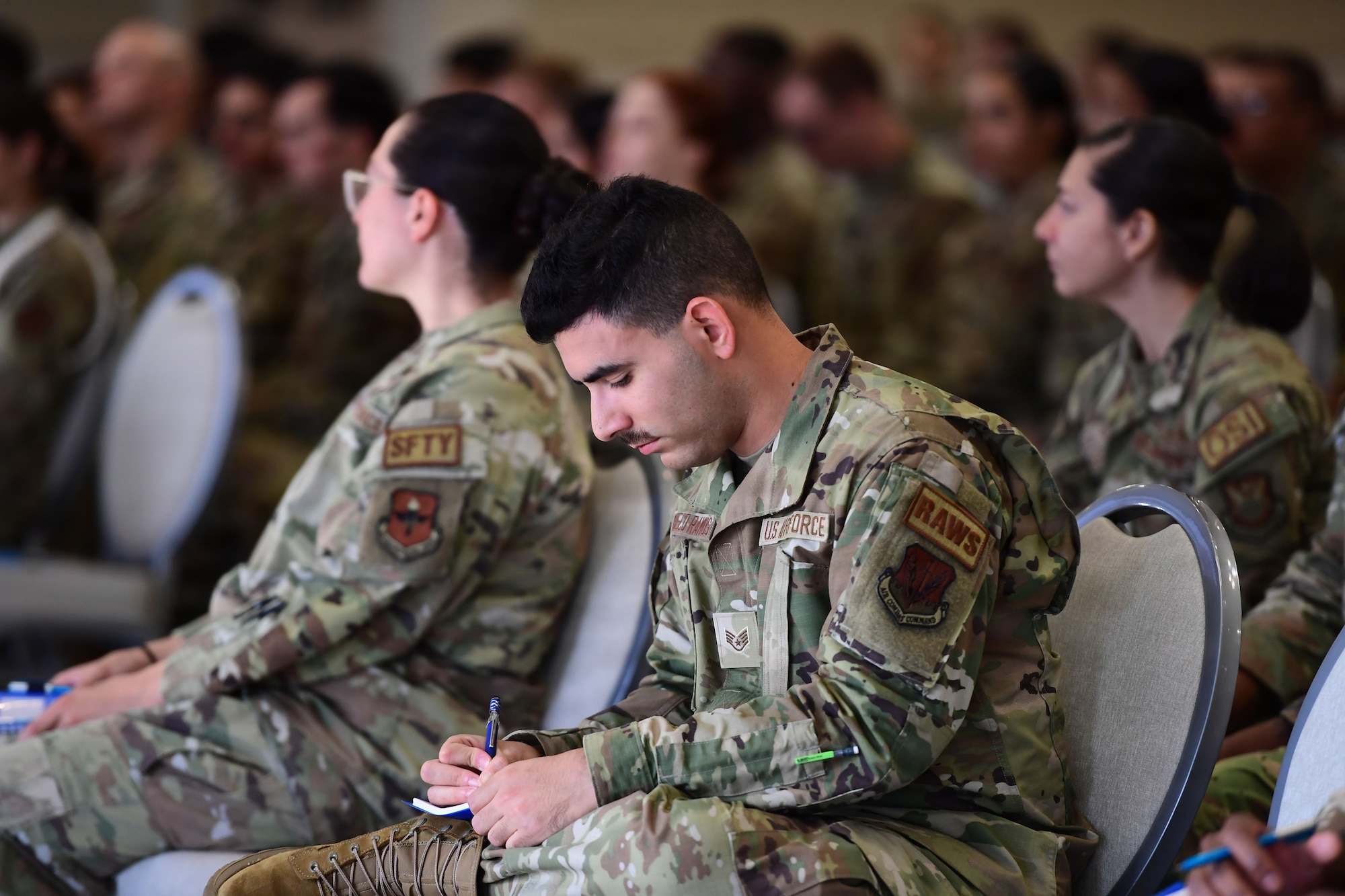 A man wearing a U.S. Air Force uniform sits in among a crowd of Airmen while taking notes.