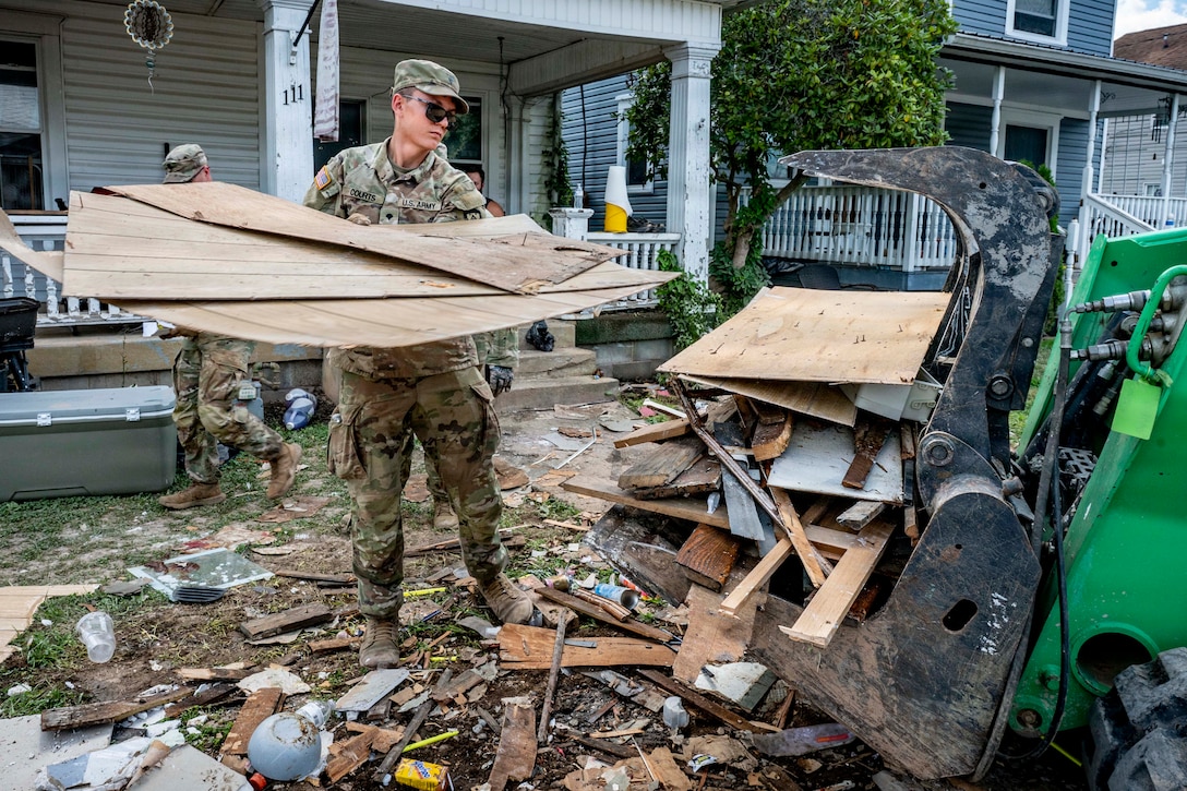 A person in a camouflage uniform in front of a house carries wooden boards toward construction equipment on a yard full of debris.