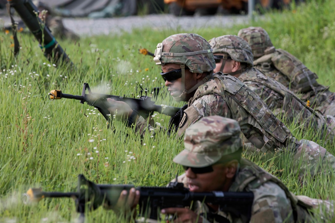 Four people in camouflage uniforms lie stomach-down in a field with their heads up and rifles pointed ahead.