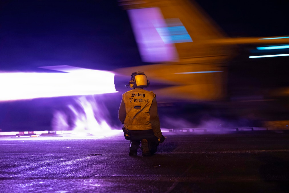 A person in a vest and helmet kneels on a flight deck at night as a jet takes off. White fire shoots out of the jet's tailpipe. The photo has a purple hue.